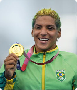 Fotografia da atleta Ana Marcela Cunha, com roupa contendo a bandeira do Brasil, segurando a medalha de ouro que está em seu pescoço. Ela está sorrindo.