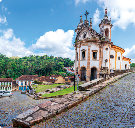 Fotografia de uma igreja com sua rua da frente e lateral feitas de paralelepípedos. A igreja tem duas torres nas laterais com uma cúpula acima. No alto de seu centro há uma cruz. Próximo a ela há várias casas e vegetação ao fundo. Em uma das ruas, alguns carros estacionados.