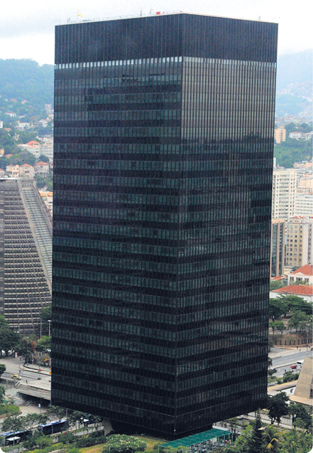 Fotografia de um edifício preto, no formato de um bloco retangular, com uma paisagem de cidade ao fundo.