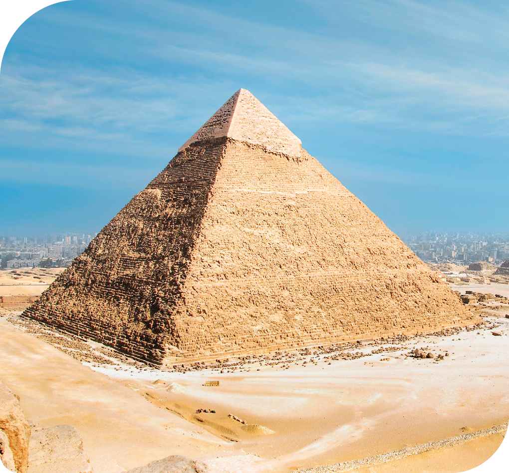 Fotografia de uma pirâmide egípcia. Ela tem a base quadrangular, está em uma área deserta com a paisagem de uma cidade ao fundo.