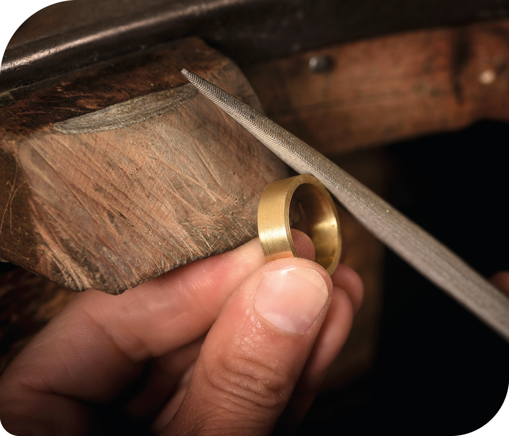 Fotografia de uma mão de uma pessoa segurando um anel de ouro, semelhante a uma aliança. Há uma lima encostada no anel, como se estivesse dando acabamento a ele.