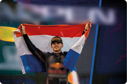 Fotografia do atleta Max Verstappen, com macacão de corrida e de boné. Ele está segurando a bandeira da Holanda atrás do corpo e com as mãos para o alto. Ele está sorrindo.