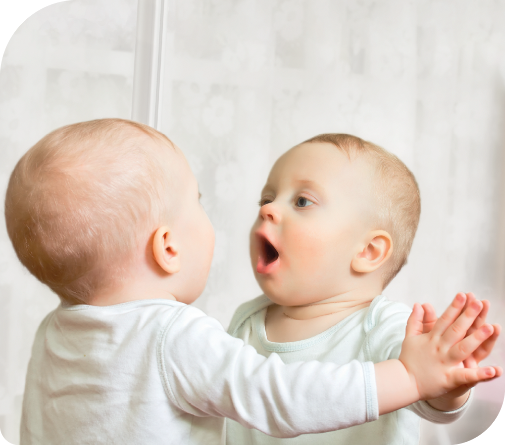 Fotografia. Um bebê com a mão apoiada em um espelho vertical, olhando seu reflexo.