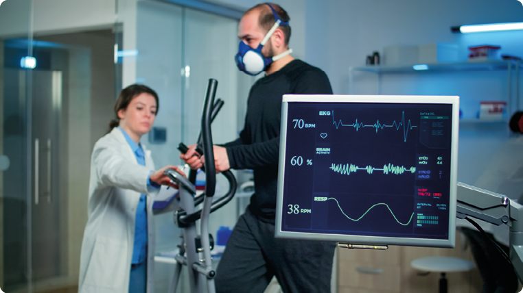 Fotografia. Um homem fazendo teste cardíaco e exames em um aparelho de atividade física, enquanto é monitorado por uma médica. Ao lado, há um monitor exibindo seus batimentos cardíacos e outros registros de monitoramento médico. 