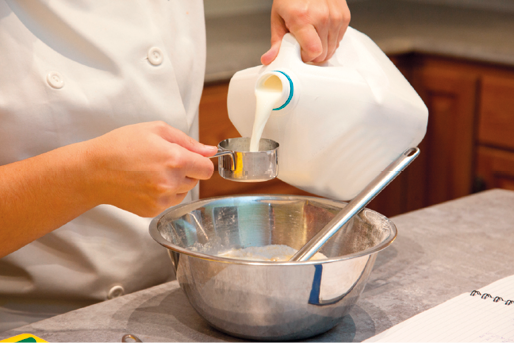 Fotografia das mãos de uma pessoa medindo a quantidade de leite diante de um preparo de alimento.