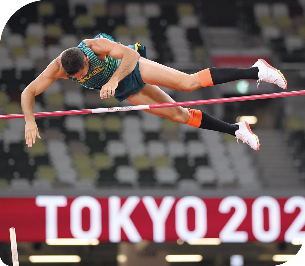 Fotografia de um atleta em pleno salto com vara. Ele está no ar, com o corpo na horizontal, barriga para baixo e  está acima de uma barra horizontal.