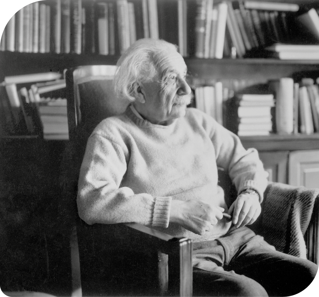 Fotografia. Um homem de bigode e cabelos brancos na altura do ombro (Albert Einstein), está sentado em uma poltrona, com os braços apoiados sob suas laterais. Ele está pensativo e de perfil na fotografia. Ao fundo, há uma estante de livros.