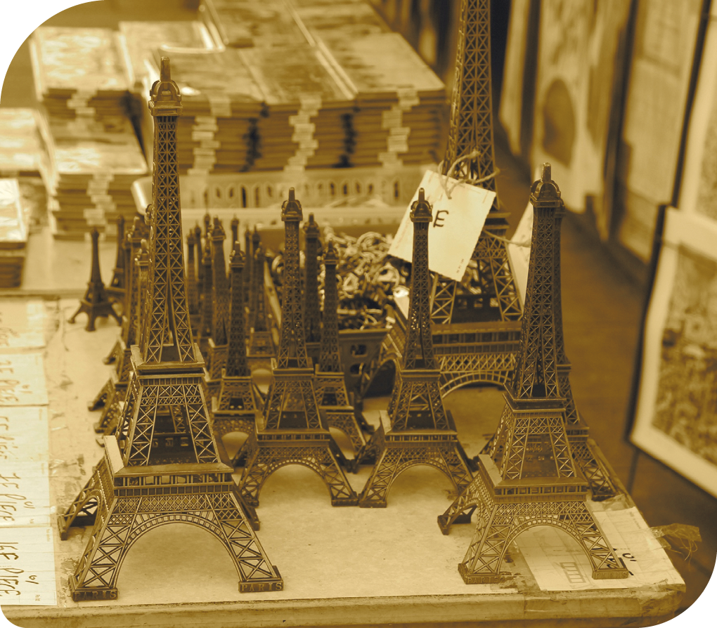 Fotografia. Várias miniaturas da Torre Eiffel de diferentes tamanhos. Estão sobre uma mesa e em tons de cobre e marrom.