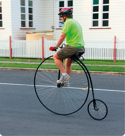 Fotografia de um homem andando com de bicicleta em uma rua de asfalto, com a roda da frente maior que a roda de trás. Ao fundo uma cerca e uma casa.