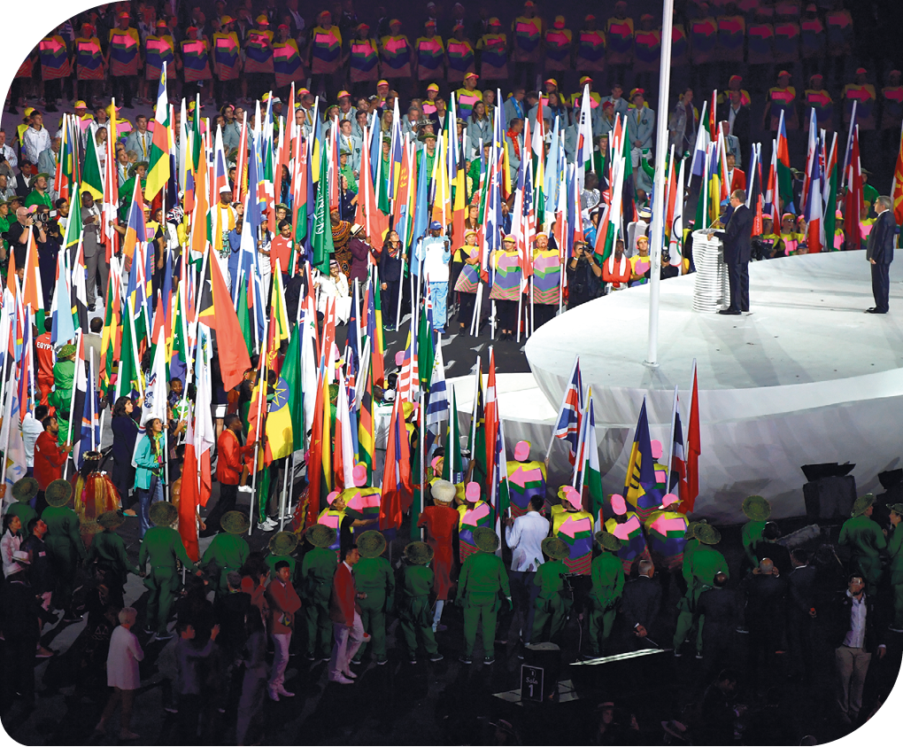 Fotografia. Cerimônia de abertura dos Jogos Olímpicos de Verão. Há um palco com um púlpito e um homem fazendo o cerimonial e em volta do palco, há vários atletas e também várias bandeiras de diferentes países, em mastros, sendo seguradas por algumas pessoas.