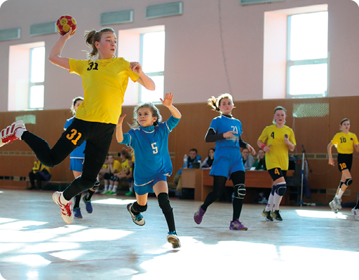 Fotografia de meninas jogando handebol na quadra de um ginásio. Uma menina está arremessando a bola. 