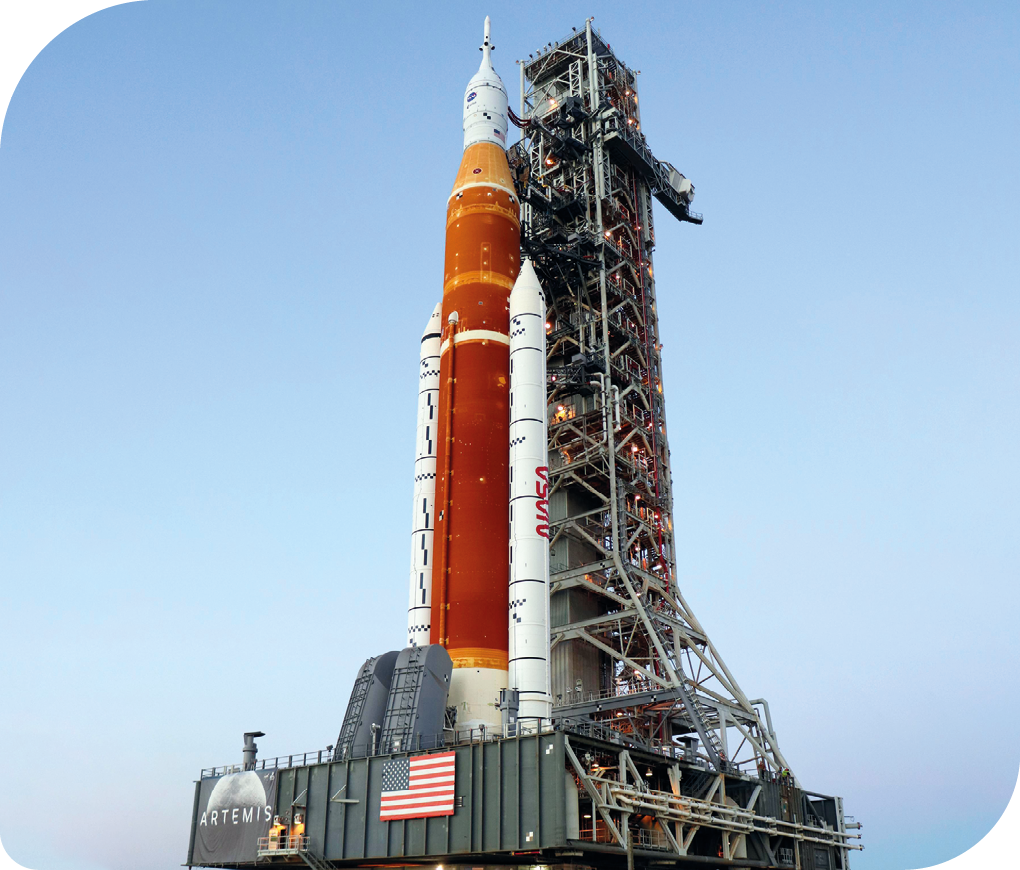 Fotografia de um foguete espacial, ao lado de uma torre de ferro vertical, e, na extremidade inferior, uma plataforma horizontal ao qual ele está apoiado