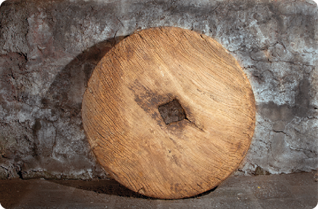 Fotografia de uma roda de madeira, com um quadrado vazado ao centro, apoiada em uma parede rochosa.