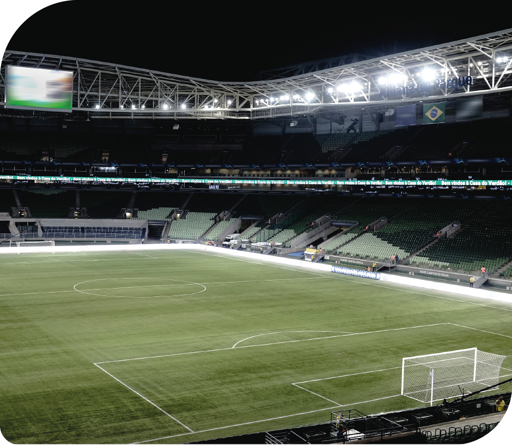 Fotografia. Estádio de futebol, com o campo de grama verde e com linhas que marcam o meio do campo, a área de gol e a limitação do campo. Há um telão e arquibancadas nas laterais. Também há luzes, e está noite.