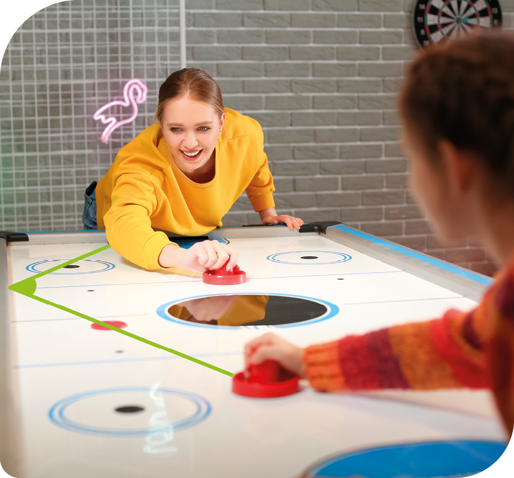 Fotografia. Duas meninas jogando Air Hockey. Há uma mesa lisa com desenhos circulares e as meninas seguram um suporte liso nas mãos que desliza sobre a mesa para tentar rebater um disco, que também desliza. Elas estão apoiadas em uma das mãos, enquanto disputam para acertar o disco. Há uma ilustração, em verde, indicando o ângulo formado pela trajetória do disco batendo na lateral da mesa. 