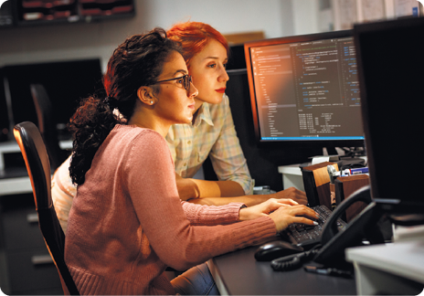 Fotografia de duas mulheres diante de um computador.  Uma delas está digitando, e a outra está apoiada na mesa olhando para a tela.
