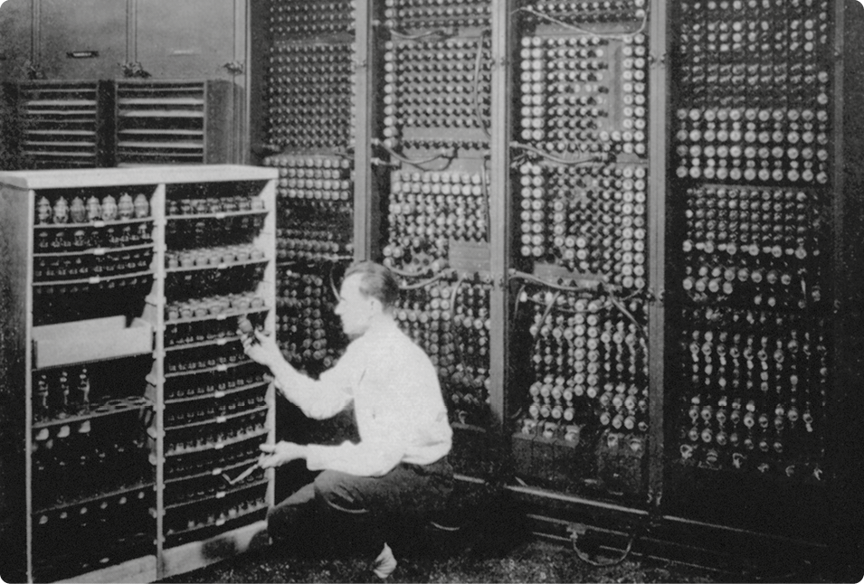 Fotografia em preto e branco de um grande computador que ocupa toda a extensão da parede. Parte desse computador também está em uma estrutura como uma estante. Há um adulto no local.