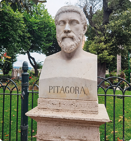Fotografia da escultura de busto de um homem de barba e bigode. Está escrito PITAGORA na própria escultura, na parte de baixo. Ao fundo há árvores e uma grade próxima.