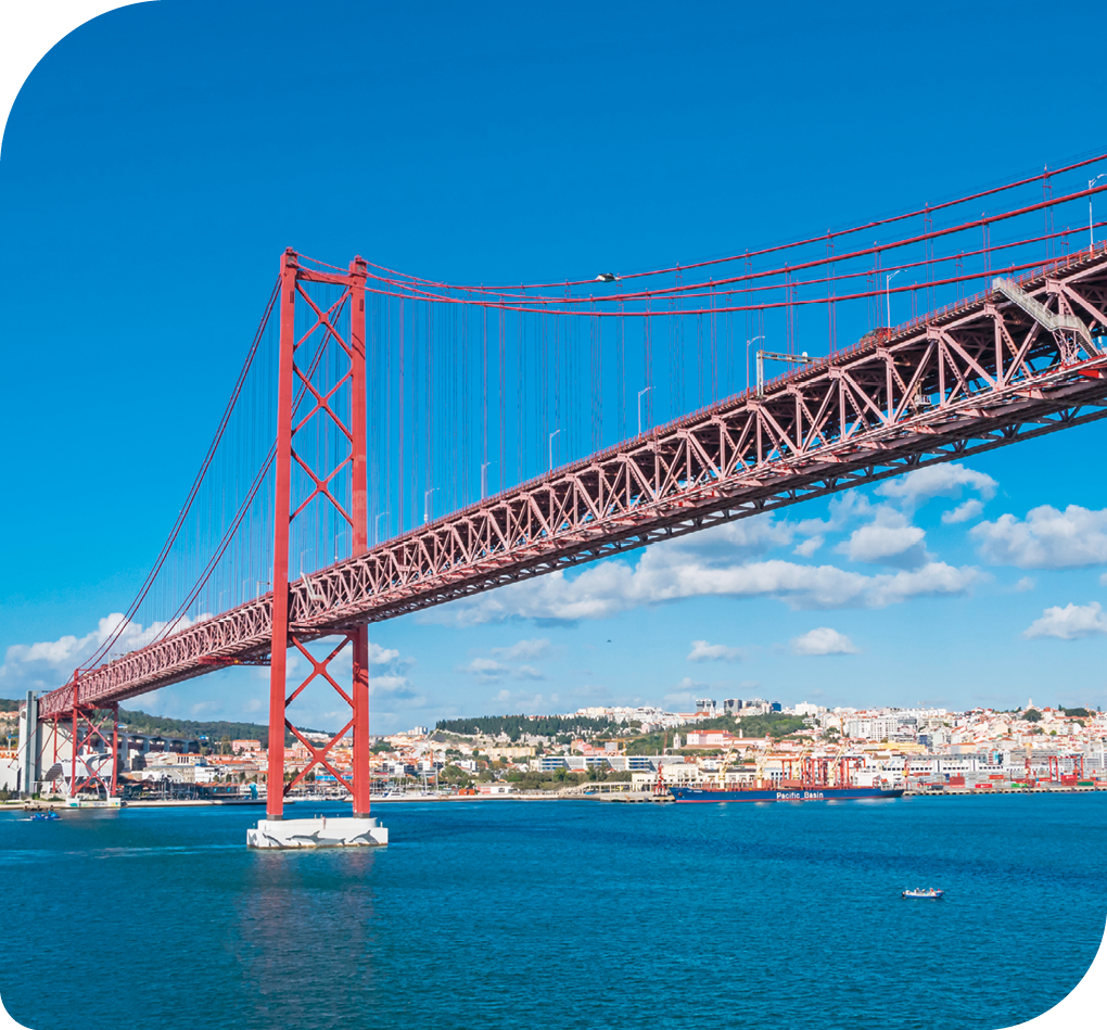 Fotografia em visão oblíqua, com a vista lateral de uma ponte pênsil sobre uma grande superfície de água. Ao fundo há uma cidade onde a ponte vai de encontro.
