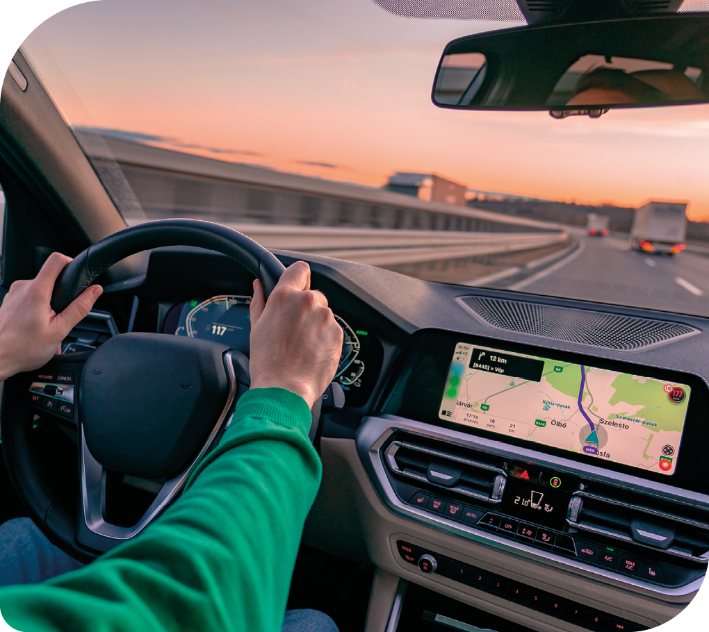 Fotografia  do interior de um carro. As duas mãos de uma pessoa estão no volante, e no painel há uma tela com um mapa aberto. O carro está em uma estrada vista a partir do para-brisa.