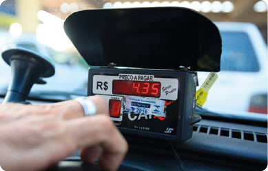 Fotografia de um taxímetro no interior de um carro. No aparelho e ao lado do visor, está indicado o símbolo R $ e no visor há o número 4,35. Acima do visor há a indicação 'PREÇO A PAGAR'.