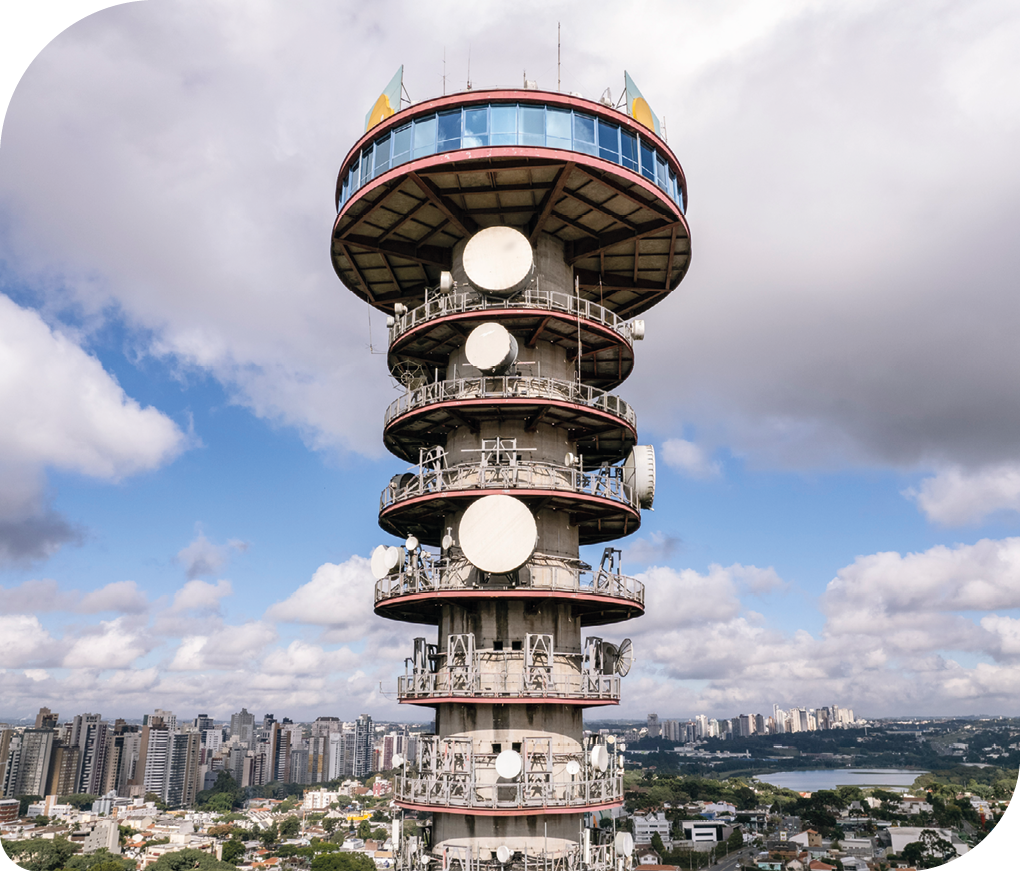 Fotografia de uma torre de telecomunicação. A torre tem um formato cilíndrico. O último andar possui um diâmetro maior que os demais andares. Há antenas que lembram círculos, distribuídas em alguns andares. Por toda a extensão da superfície, na parte debaixo da foto, há uma cidade e há um lago ao fundo e à direita.