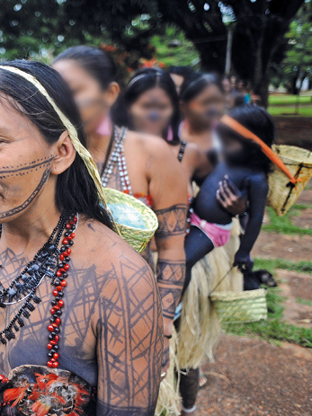 Fotografia. Vista parcial de mulheres indígenas em fila, com pinturas em preto pelo corpo, colares com miçangas em preto, vermelho e branco, cabelos longos pretos. À frente, a mulher traz sobre a cabeça a alça de um pequeno cesto de palha que se apoia em suas costas, na altura do ombro esquerdo. Uma das mulheres traz uma criança de aproximadamente 2 anos no colo. Essa criança está com o corpo pintado de preto e também traz na cabeça a alça de um pequeno cesto de palha que se apoia em suas costas, na altura da nuca. Ao fundo, vê-se o local com solo marrom e vegetação rasteira em verde.