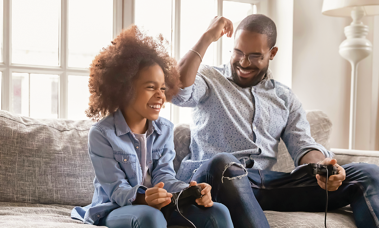 Fotografia. Homem com barba, camisa branca e calças jeans, está sentado num sofá cinza junto de uma menina de cabelos armados, usando camisa azul-clara e calças jeans. Ambos sorridentes jogando videogame. O homem está com o braço direito levantado em gesto de comemoração. Ao fundo uma vidraça, do lado direito do sofá, um abajur branco.