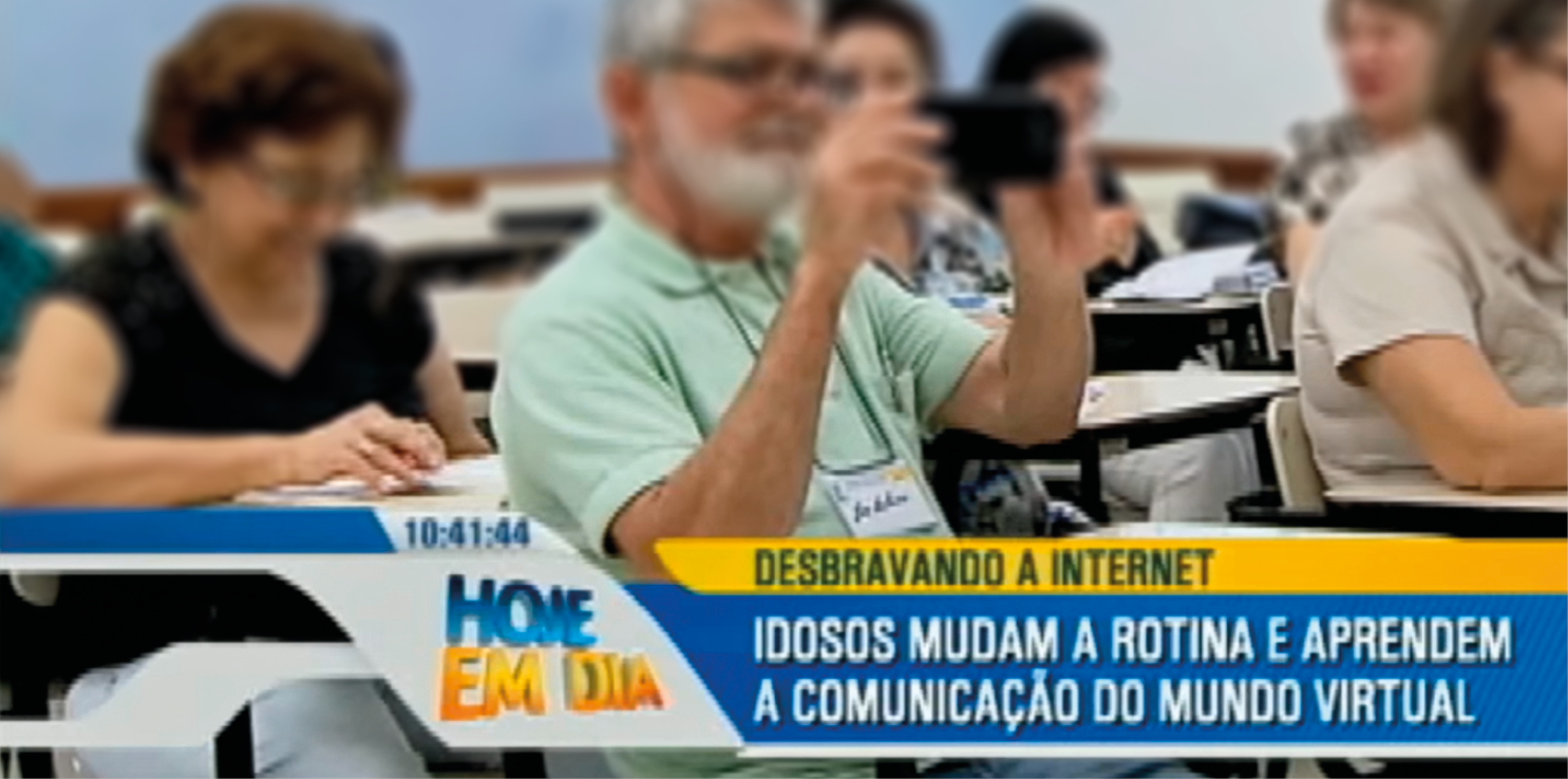 Captura de tela. Em uma sala de aula, pessoas idosas sentadas em carteiras escolares. Ao centro, um homem de cabelos brancos com camiseta e gola em verde-claro, com par de óculos de grau, segurando nas mãos um celular preto na horizontal. Na parte inferior, texto: 10:41:44 HOJE EM DIA. Desbravando a internet – Idosos mudam a rotina e aprendem a comunicação do mundo virtual.