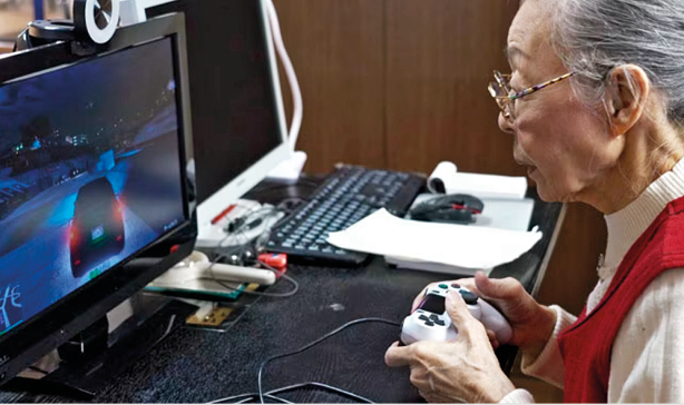 Fotografia. À direita, uma pessoa de frente para mesa com computador preto, jogando videogame. Ela é uma senhora de cabelos grisalhos penteados para trás, blusa branca e colete vermelho por cima, com par de óculos de grau.