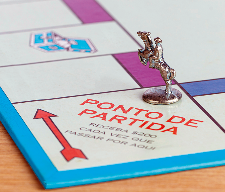 Fotografia. Vista acima de mesa de madeira com foco de um jogo de tabuleiro. Ele tem a cor branca com detalhes na borda em azul-claro, com vários quadrados em branco. Detalhe em um quadrado onde está escrito: PONTO DE PARTIDA. Flecha para a esquerda.