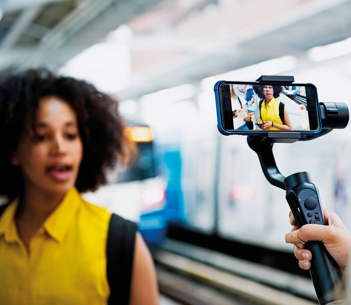 Fotografia. Uma pessoa segurando um suporte na vertical em preto onde há acoplado um celular na horizontal. Pelo celular, vê-se a imagem de uma mulher à frente. Ela tem cabelos crespos escuros, usando uma blusa regata de gola em amarelo, com detalhes nas mangas em preto. Ela olha para frente, com a boca um pouco aberta. Ao fundo, um automóvel passando visto parcialmente em branco e azul.