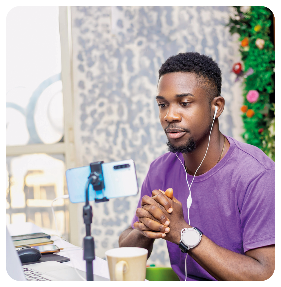 Fotografia. Um homem sentado, de frente para uma mesa branca com notebook cinza e perto, um celular na horizontal sobre um pedestal. O homem tem cabelos, bigode e barba escuros, de camiseta em roxo, com um fone de ouvido e relógio no braço esquerdo, com as mãos entrelaçadas. Ao fundo, vista parcial de parede em azul-claro e branco e mais à direita, folhas verdes.