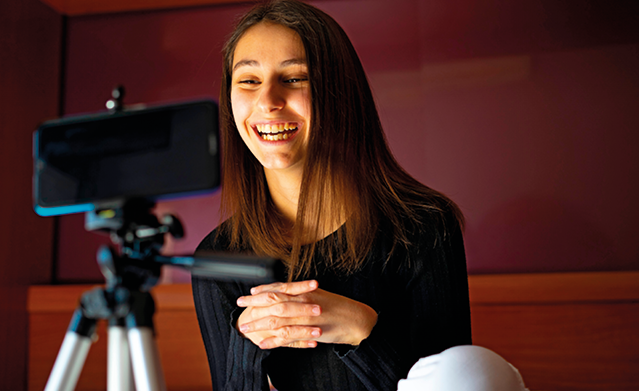 Fotografia. Uma menina vista da cintura para cima, de cabelos longos castanhos com blusa de mangas compridas em preto. Ela olha para frente sorrindo, de frente para um celular na horizontal sobre um pedestal.