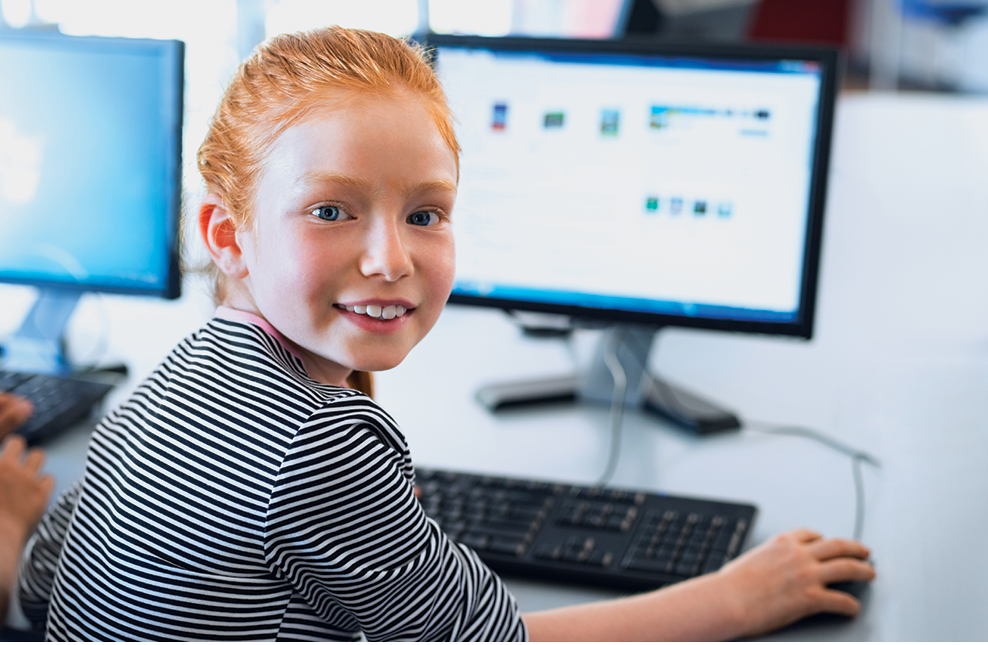 Fotografia. Uma menina vista dos ombros para cima, sentada de frente para um computador, olhando para trás. Ela tem cabelos ruivos penteados para trás, usando blusa listrada em branco e preto, segurando um mouse com a mão direita. Ela olha sorrindo.
