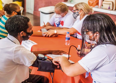 Fotografia. Um grupo de cinco pessoas sentadas de frente para uma mesa com toalhas de cor laranja. Todas usam máscara tapando o nariz e a boca. À frente, um menino com camiseta de gola alta, branca e à direita, mulher de cabelos escuros até os ombros e camiseta branca.