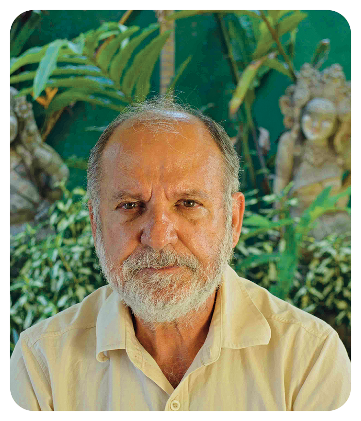 Fotografia. Um homem visto dos ombros para cima, com rugas, calvo na frente, com cabelos, bigode e barba grisalhos, usando camisa de gola em amarelo-claro. Ele olha para frente com leve sorrindo. Ao fundo, vegetação de folhas em verde.