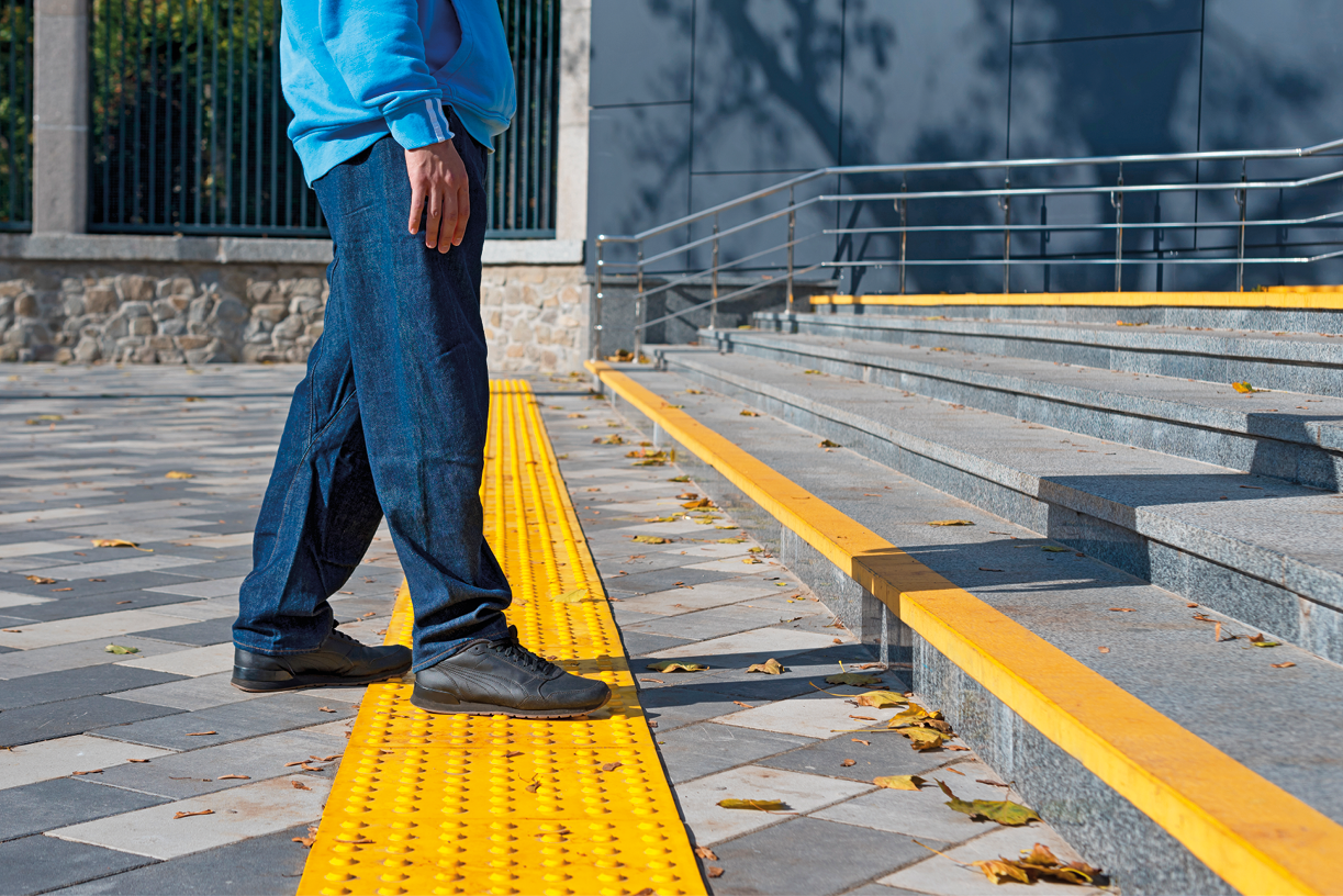 Fotografia. Vista geral de local com escada em cinza-claro e faixa na horizontal em amarelo. À esquerda, faixa com protuberâncias por onde passa um homem caminhando para a direita. Ele veste camisa em azul-claro, de calça jeans e sapatos em preto. Em segundo plano, corrimão em cinza.