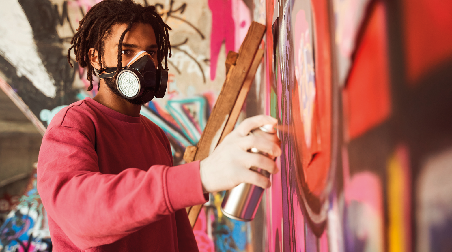 Fotografia. Um homem visto da cintura para cima pintando um muro com um tubo cinza de spray. Ele tem cabelos pretos trançados, de blusa de mangas compridas em vermelho. No muro, tinta em rosa, preto e vermelho.