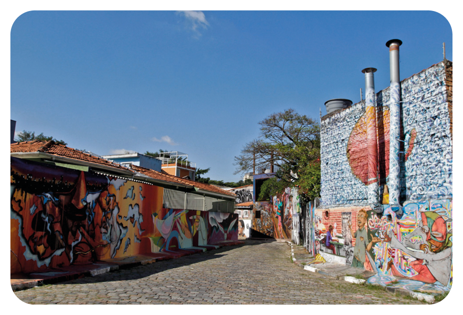 Fotografia. Uma viela, com paredes à esquerda e à direita, cobertas por grafites coloridos. À esquerda, pinturas em tons de vermelho, amarelo e laranja e à direita, em tons de cinza e vermelho. Ao fundo, casas, árvores de folhas verdes e céu em azul-claro.