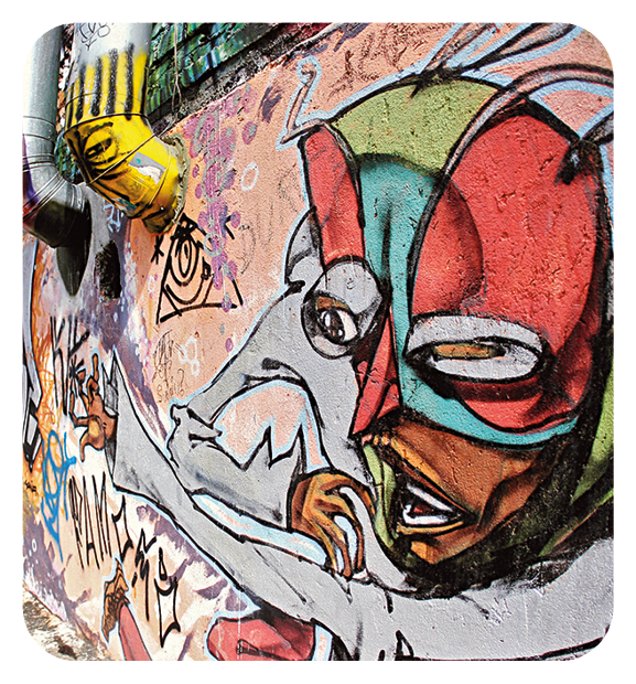 Fotografia. Detalhe de muro com ilustração de rosto de um homem e, acima do rosto, máscara vermelha com detalhes em azul-claro, do nariz para cima. Ele usa blusa de mangas compridas em cinza e mão direita sobre a boca. Ao fundo, partes do muro em rosa-claro.