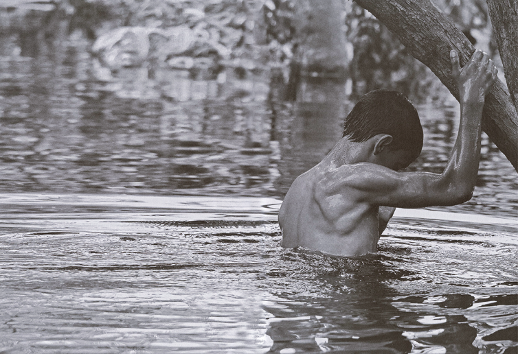 Fotografia em preto e branco. Um jovem visto de costas, em um rio, com o corpo da cintura para cima, braço direito segurando em ramo de árvore. Ele tem cabelos escuros lisos, músculos flexionados do braço. Em segundo plano, rio e reflexo da vegetação ao redor.
