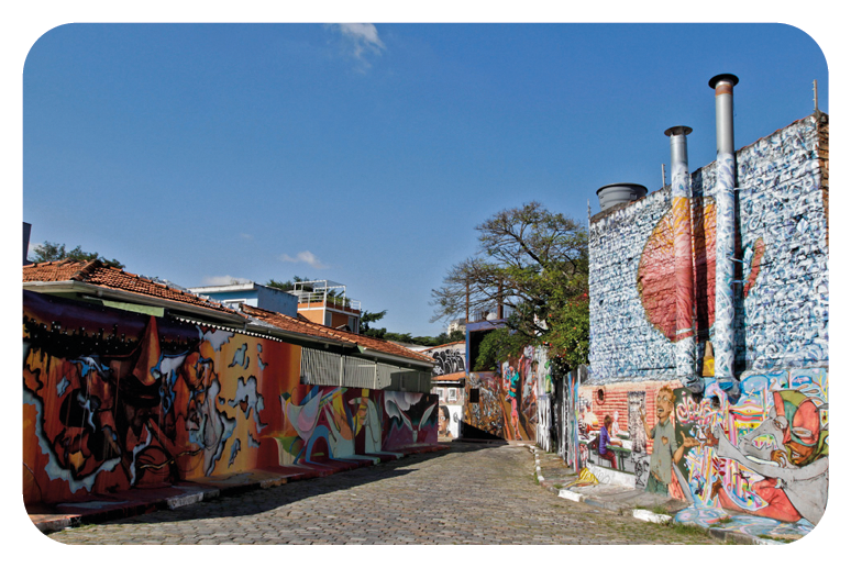 Fotografia. Uma viela, com paredes à esquerda e à direita, cobertas por grafites coloridos. À esquerda, pinturas em tons de vermelho, amarelo e laranja e à direita, em tons de cinza e vermelho. Ao fundo, casas, árvores de folhas verdes e céu em azul-claro.