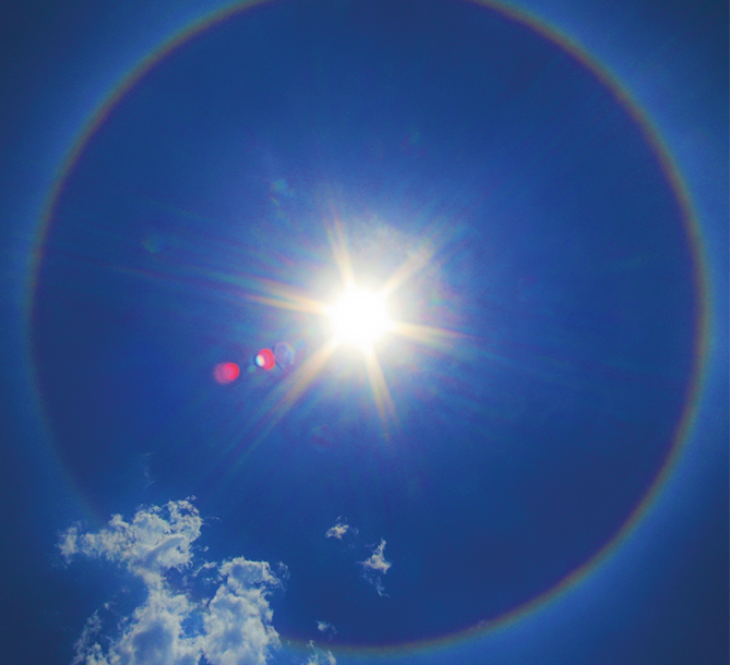 Fotografia. Vista do alto de um céu em azul-escuro. Um círculo em azul com contorno em preto. Ao centro, um sol irradiante e abaixo, nuvens brancas.