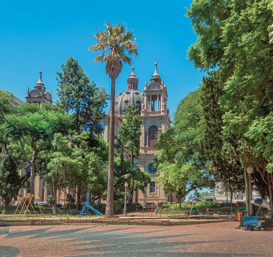 Fotografia. Vista geral de local aberto. À frente, rua em cinza com partes em marrom. Mais ao fundo, árvores de folhas verdes, algumas médias e outras altas. Em segundo plano, vista parcial de uma igreja, com torres em marrom-claro e uma cúpula ao centro. No alto, céu em azul-claro, sem nuvens.