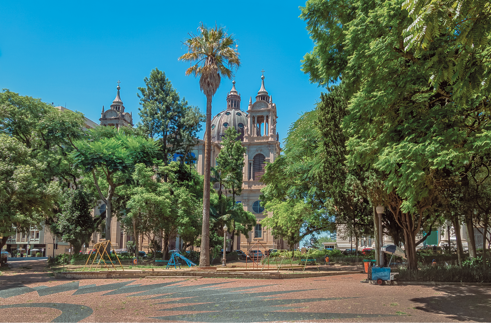 Fotografia. Vista geral de local aberto. À frente, rua em cinza com partes em marrom. Mais ao fundo, árvores de folhas verdes, algumas médias e outras altas. Em segundo plano, vista parcial de uma igreja, com torres em marrom-claro e uma cúpula ao centro. No alto, céu em azul-claro, sem nuvens.