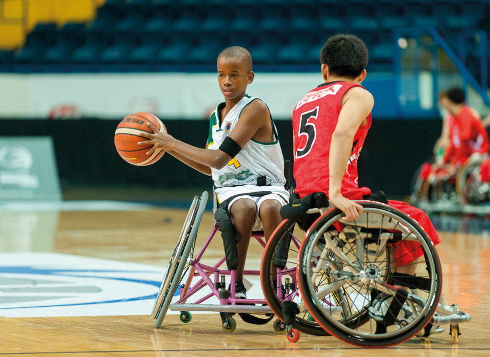 Fotografia. Em uma quadra de basquete de solo marrom, com detalhes em branco e linhas em azul. Dois jovens cadeirantes, um ao lado em jogada de basquete. À esquerda, jovem com regata branca e segurando nas mãos uma bola de basquete em laranja. À direita, um jovem visto de costas, de cabelos pretos, de regata em vermelho. Ao fundo, outros jogadores vistos parcialmente e mais ao fundo, arquibancada.