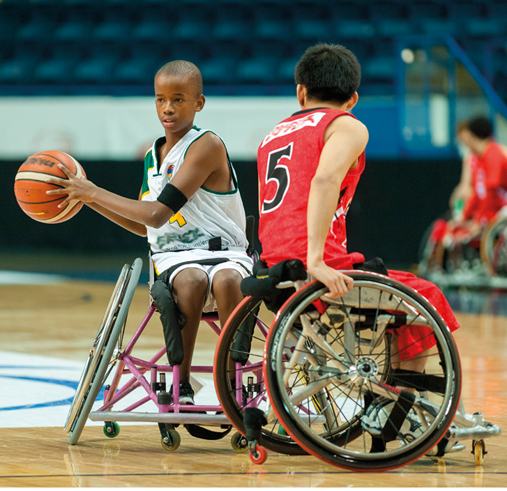 Fotografia. Em uma quadra de basquete de solo marrom, com detalhes em branco e linhas em azul. Dois jovens cadeirantes, um ao lado em jogada de basquete. À esquerda, jovem com regata branca e segurando nas mãos uma bola de basquete em laranja. À direita, um jovem visto de costas, de cabelos pretos, de regata em vermelho. Ao fundo, outros jogadores vistos parcialmente e mais ao fundo, arquibancada.