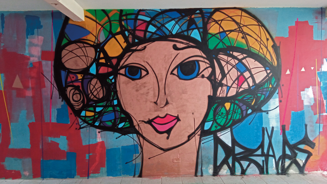 Grafite. Vista geral de um muro em azul, com pintura ao centro. Uma cabeça de pessoa de olhos azuis, lábios em rosa e sobre a cabeça, cabelos coloridos em azul, verde e laranja. À esquerda e à direita, pinturas em vermelho e à direita, a palavra DREADS pichada em preto.