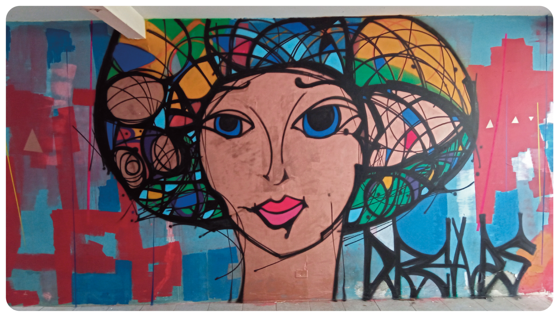 Grafite. Vista geral de um muro em azul, com pintura ao centro. Uma cabeça de pessoa de olhos azuis, lábios em rosa e sobre a cabeça, cabelos coloridos em azul, verde e laranja. À esquerda e à direita, pinturas em vermelho e à direita, a palavra DREADS pichada em preto.